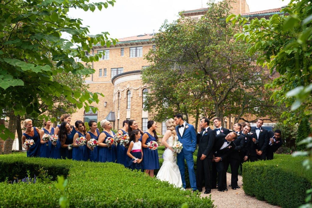 Huge bridal parties looking at bride and groom kiss each other in the private English style garden at Hotel Baker Saint Charles Illinois.