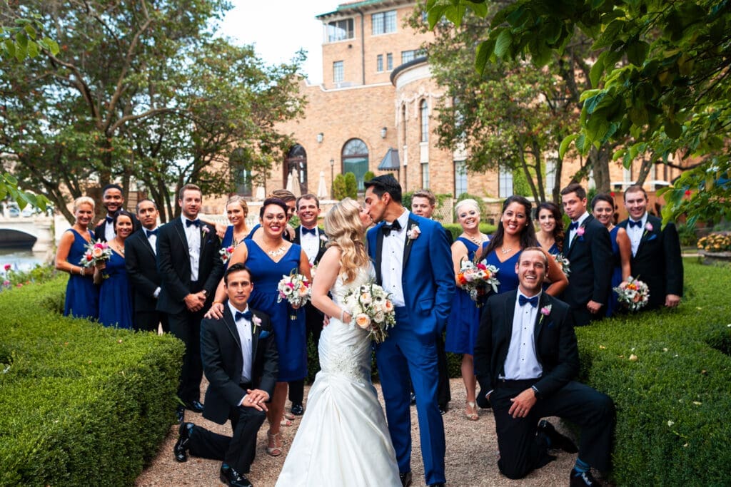 Modern bridal party looking at camera while bride and groom kiss in the courtyard at Hotel Baker.