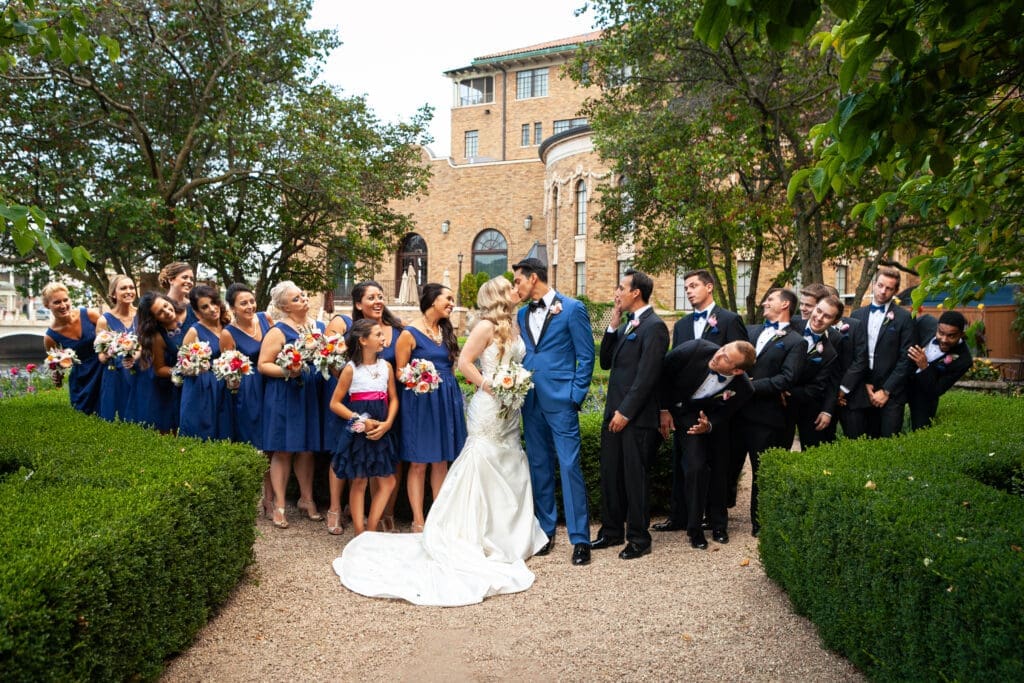 Huge bridal party look at bride and groom while they share a kiss together at Hotel Baker private garden.