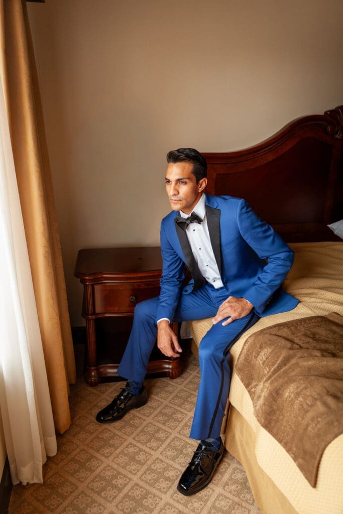 Handsome Groom sits in a hotel room at Hotel Baker awaiting his bride staring out the window in a blue tuxedo.