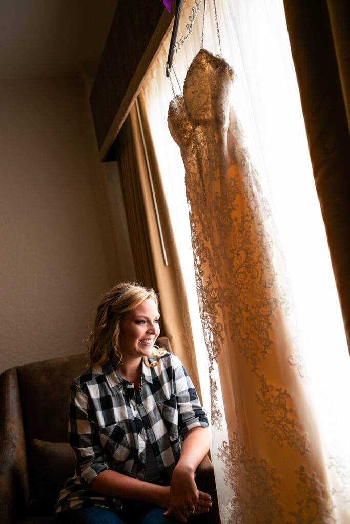 Bride sitting by her hanging wedding dress as it hangs in front of the window in the bridal suite in Hotel Baker.