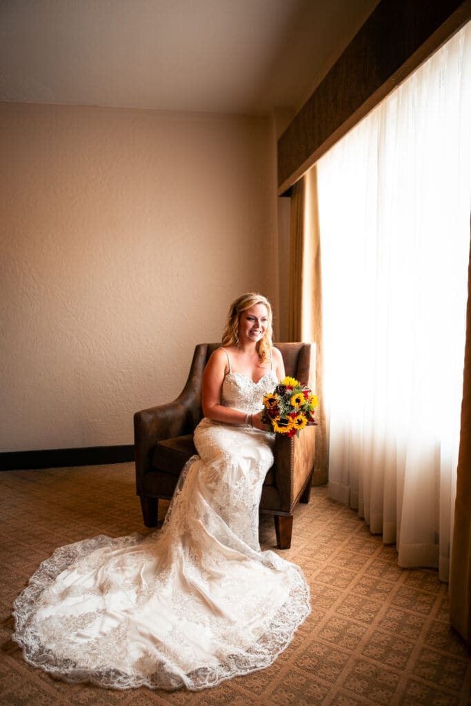 Bride sitting in chair holding her flowers as she looks out the window on wedding day.