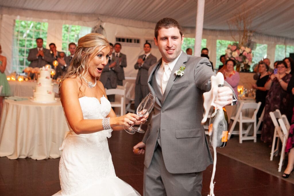 The groom holds an erupting champaign bottle as the bride looks on in shock and amazement as the champaign comes shooting out of the bottle.