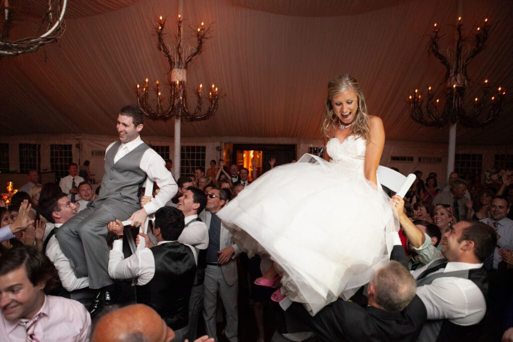 Bride and groom holding on the dancefloor smiling as they are lifting high up on chairs by their guests with chandeliers in the background look at each other.