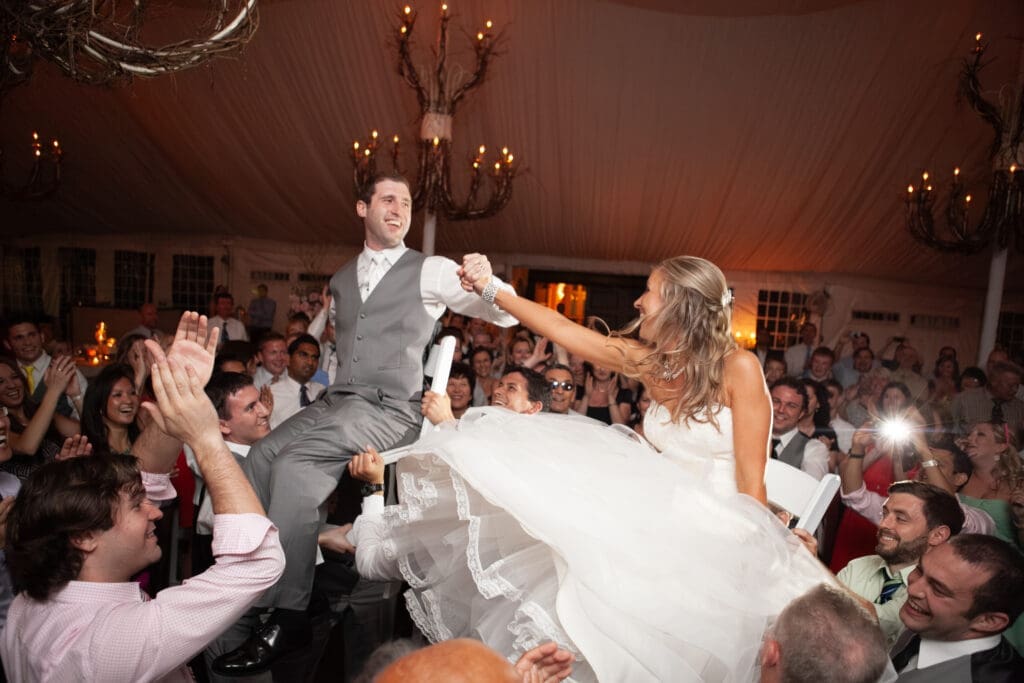 Bride and groom holding hands on the dancefloor smiling as they are lifting high up on chairs with chandeliers in the background look at each other.