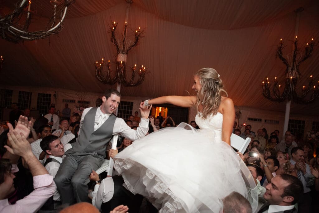 Bride and groom holding hands on the dancefloor smiling as they are lifting high up on chairs with chandeliers in the background look at each other.