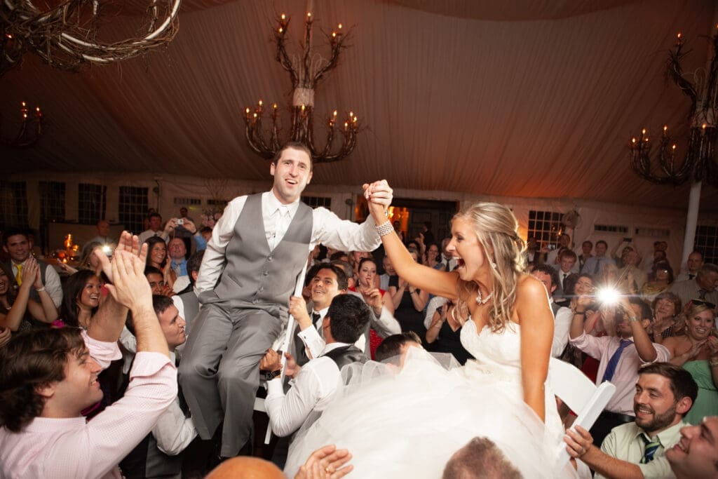 Bride and groom holding hands on the dancefloor smiling as they are lifting high up on chairs with chandeliers in the background.