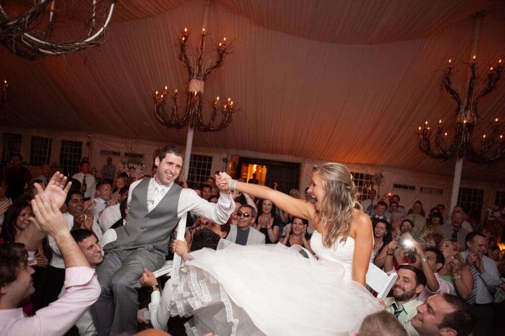 Bride and groom holding hands on the dancefloor smiling as they are lifting high up on chairs with chandeliers in the background look at each other excitedly.