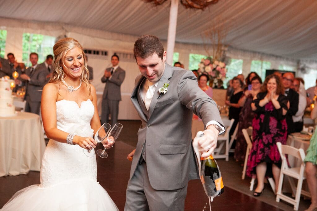 Groom holds a spilling champaign bottle as the bride holds two glasses ready to pour and enjoy.
