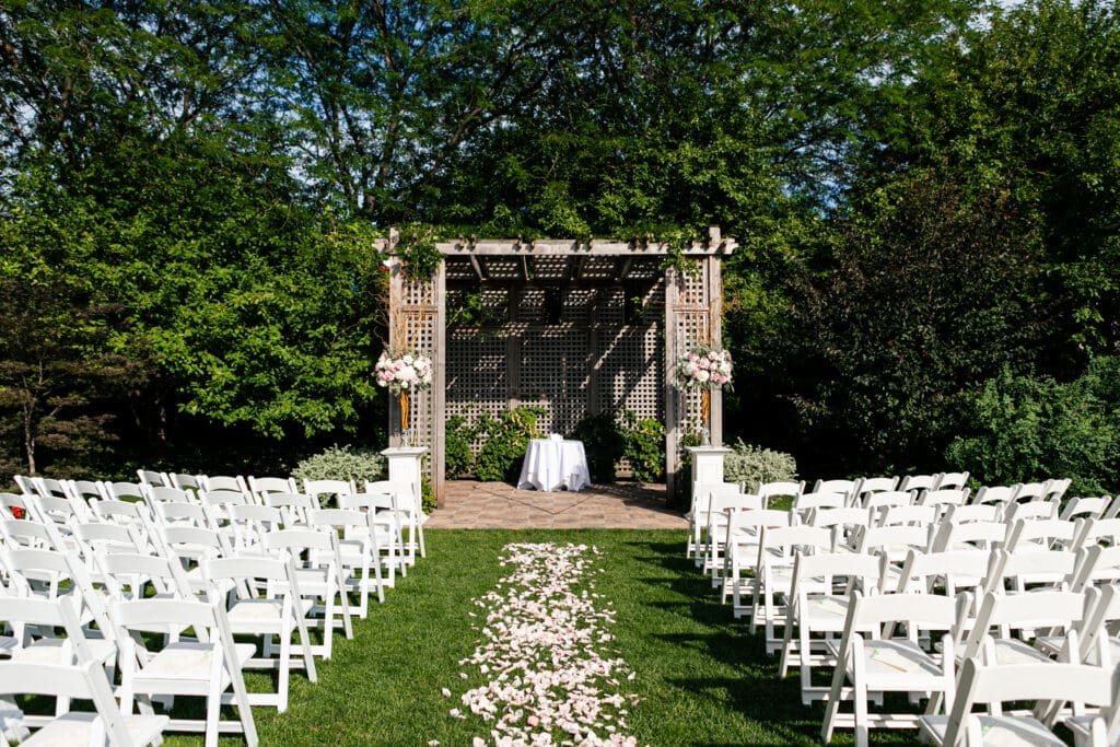 Private estate wedding ceremony site decorated with chairs, flowers on stands and rose petals down the entire aisle.